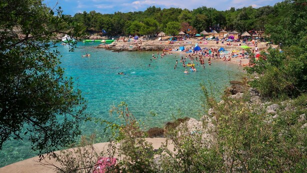 Een strand met helder turquoise water, mensen die zwemmen en zonnebaden, en een rotsachtige kustlijn met bomen op de achtergrond.