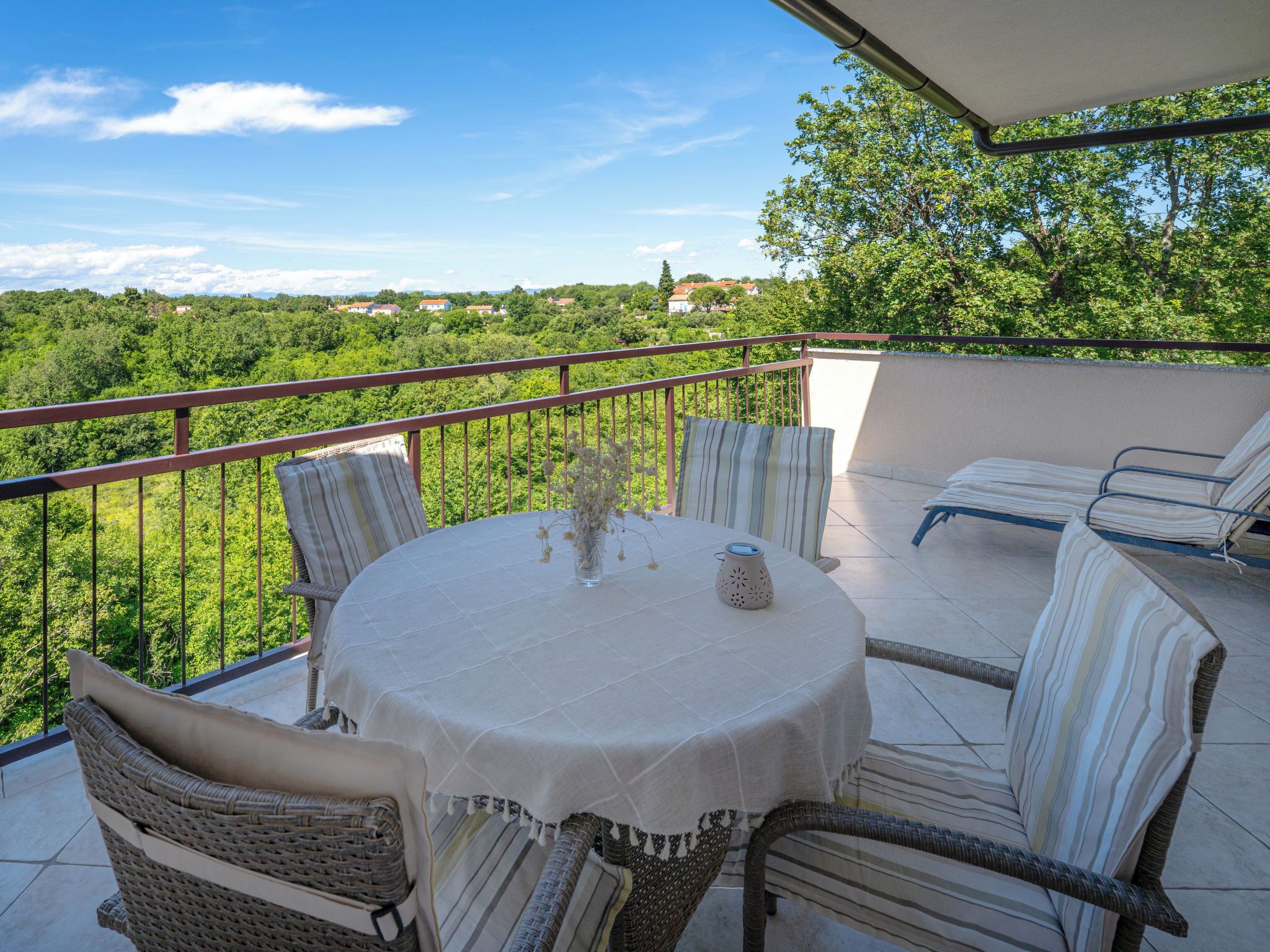 A covered outdoor seating area with a round table, wicker chairs, and a view of greenery and buildings.