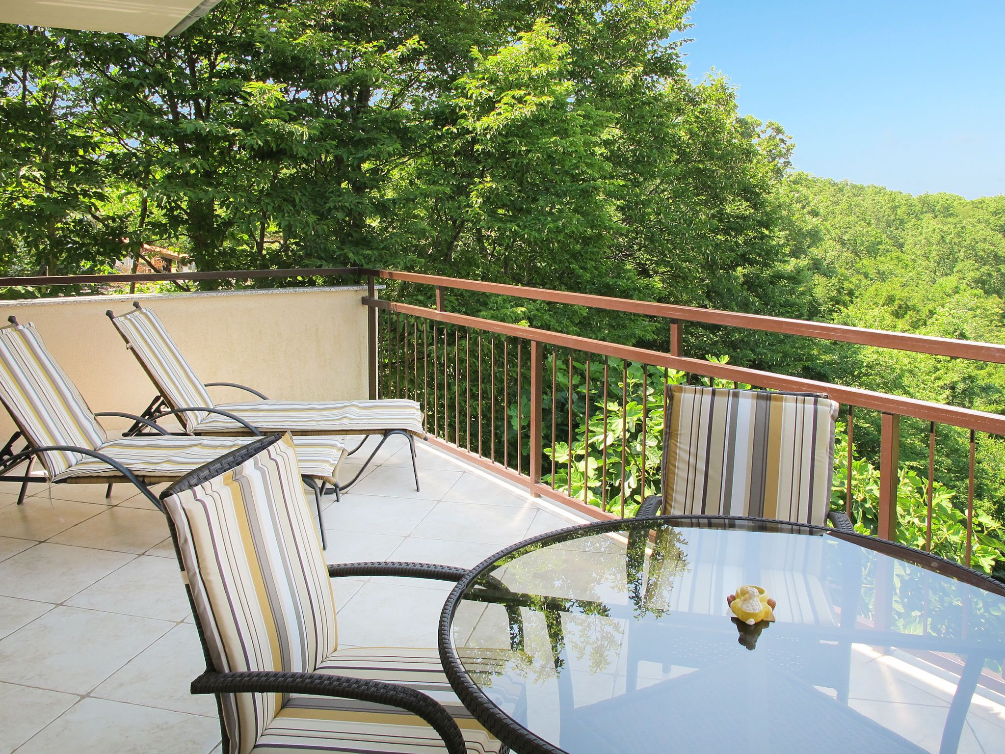 A balcony with two striped lounge chairs and a glass table, surrounded by lush greenery.