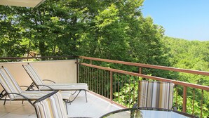 Un balcon agrémenté de deux fauteuils de jardin à rayures et d’une table en verre, le tout entouré d’une végétation luxuriante.