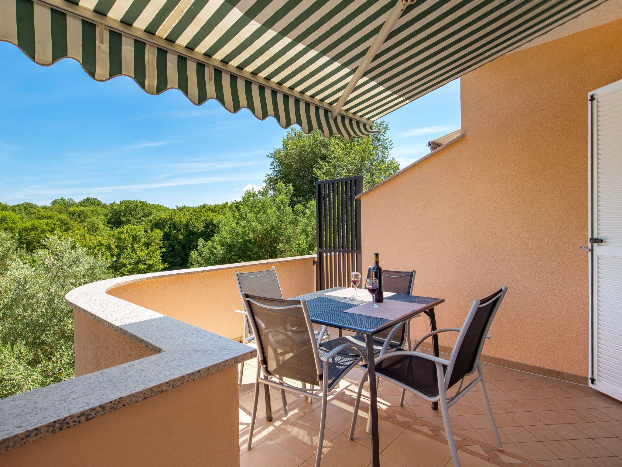 A covered outdoor seating area with a table and chairs, a bottle of wine, and a view of greenery.