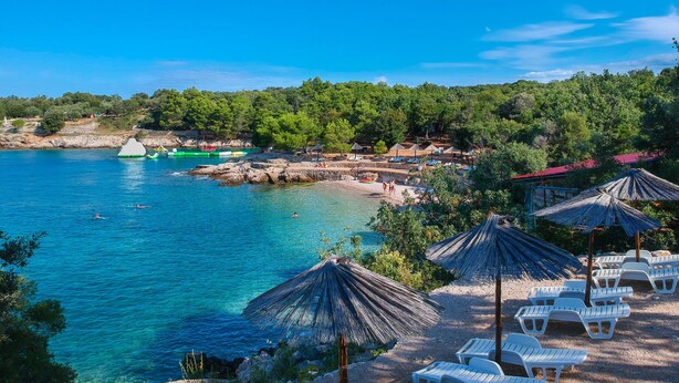 Ein Küstenstrand mit klarem, blauem Wasser, Strandzelten und Liegestühlen.