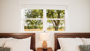 A bedroom with two beds, a nightstand with a lamp, and a window with a view of trees and fields.