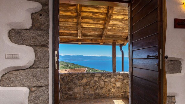Une pièce aux murs de pierre, dotée d’une porte en bois qui donne sur un balcon offrant une vue panoramique sur la mer et les montagnes.