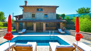 Un espace piscine avec des chaises longues et des parasols, un bâtiment en pierre avec un toit en tuiles et un ciel d’un bleu limpide.