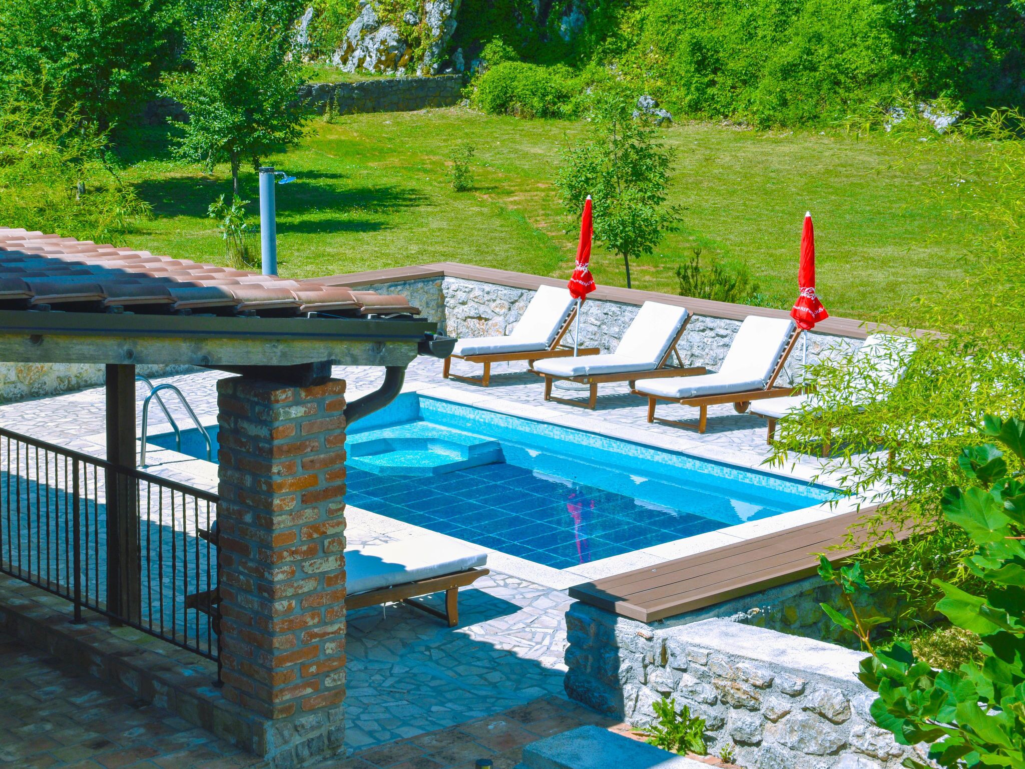Una piscina con sillas de descanso y sombrillas, rodeada de muros de piedra y vegetación.