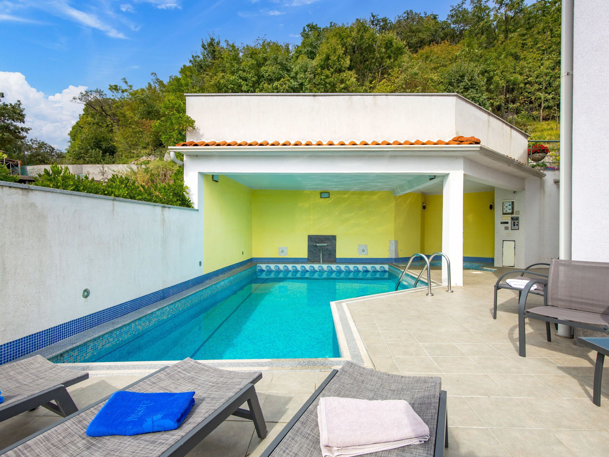 A swimming pool with a tiled deck, lounge chairs, and a covered area with yellow walls.