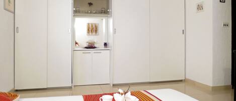 A hotel room with a bed, a breakfast setup with cups and a vase, and a minibar with glassware and a decorative item.