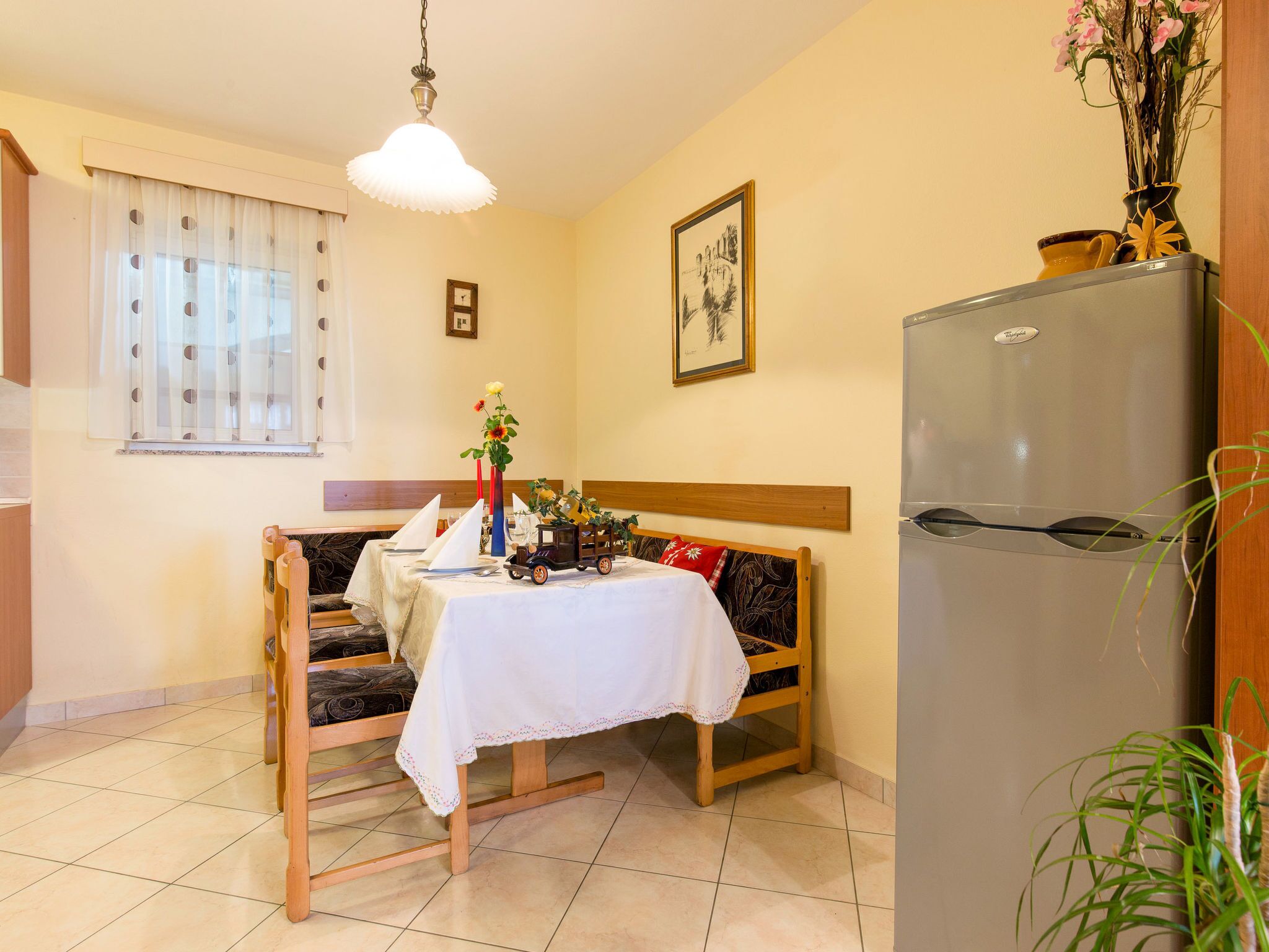 A dining area with a wooden bench, a table with a white tablecloth, a refrigerator, and a window with curtains.