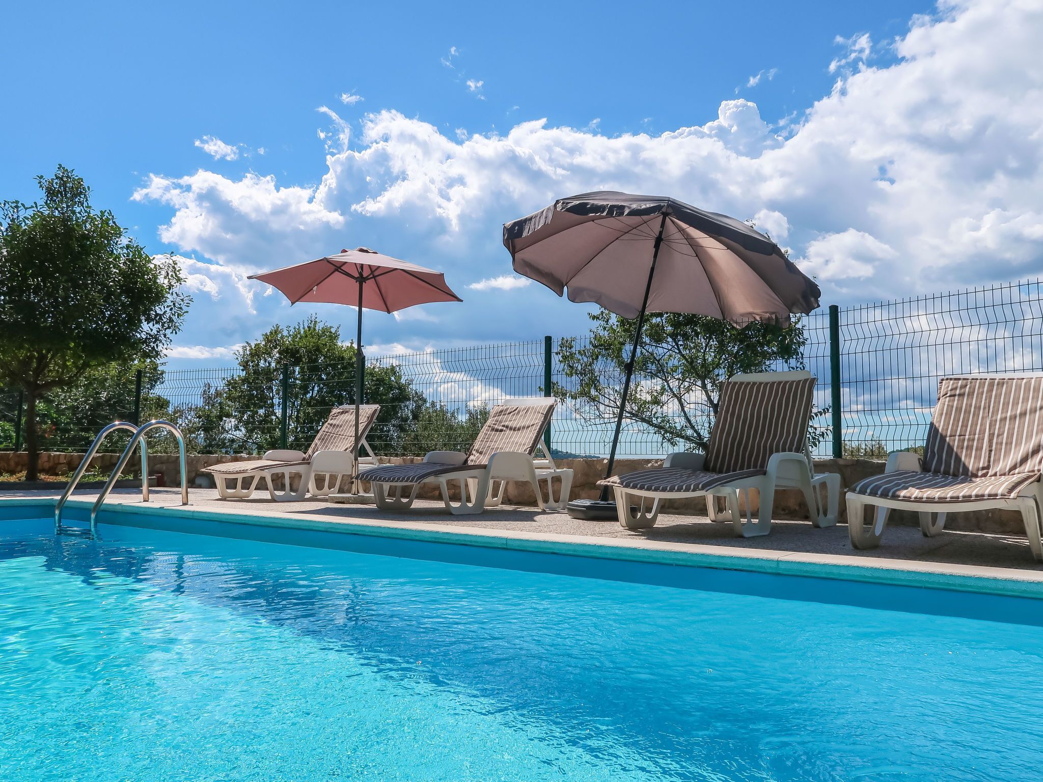 A swimming pool with lounge chairs and umbrellas, surrounded by a fence and trees.