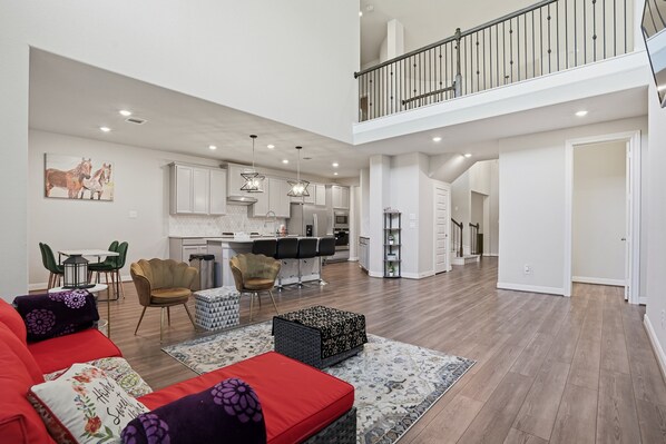 A spacious living area with a red sofa, patterned ottoman, and a dining table with chairs. A kitchen with white cabinets and a balcony is visible in the background.