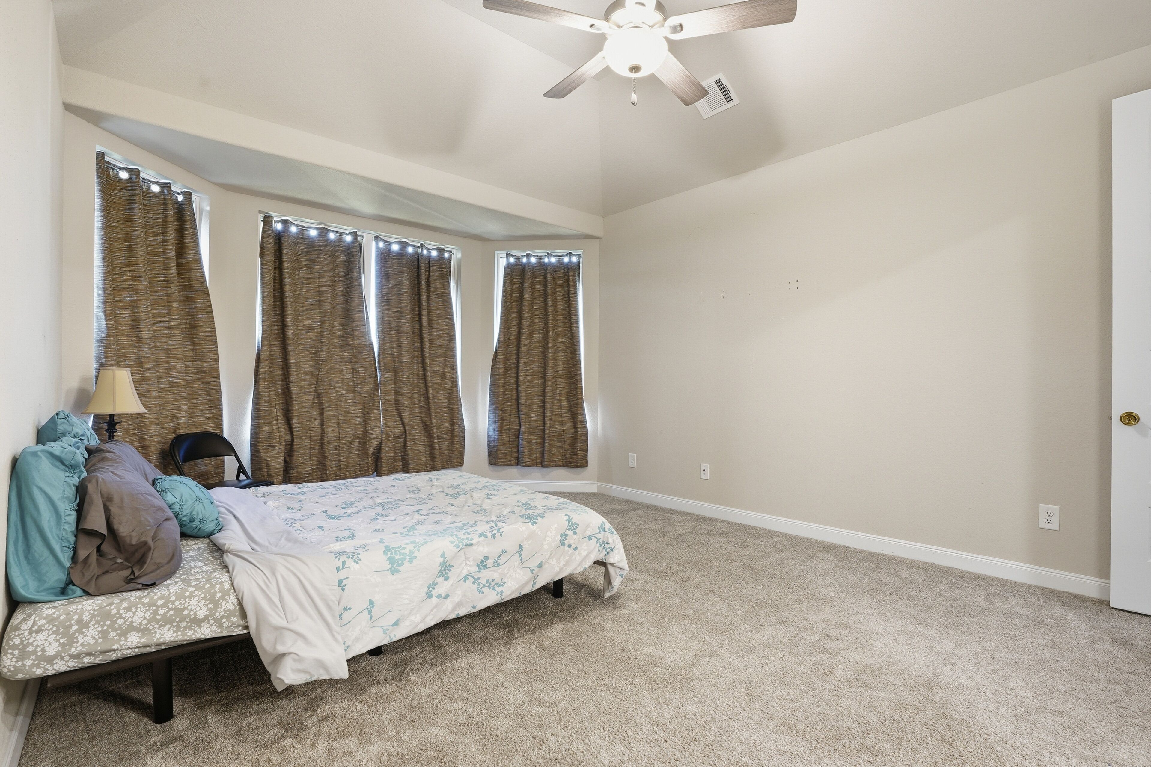 Un dormitorio con cama, ventilador de techo y tres ventanas con cortinas.