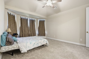 A bedroom with a bed, a ceiling fan, and three windows with curtains.
