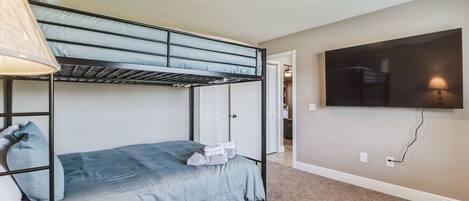 A bunk bed room with a TV mounted on the wall, carpeted floor, and a door leading to another room.
