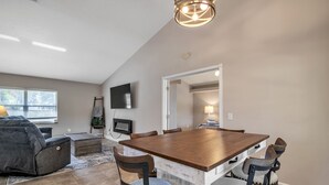 Un comedor con una mesa de madera y cuatro sillas, un televisor montado en la pared, y un ventanal con persianas.