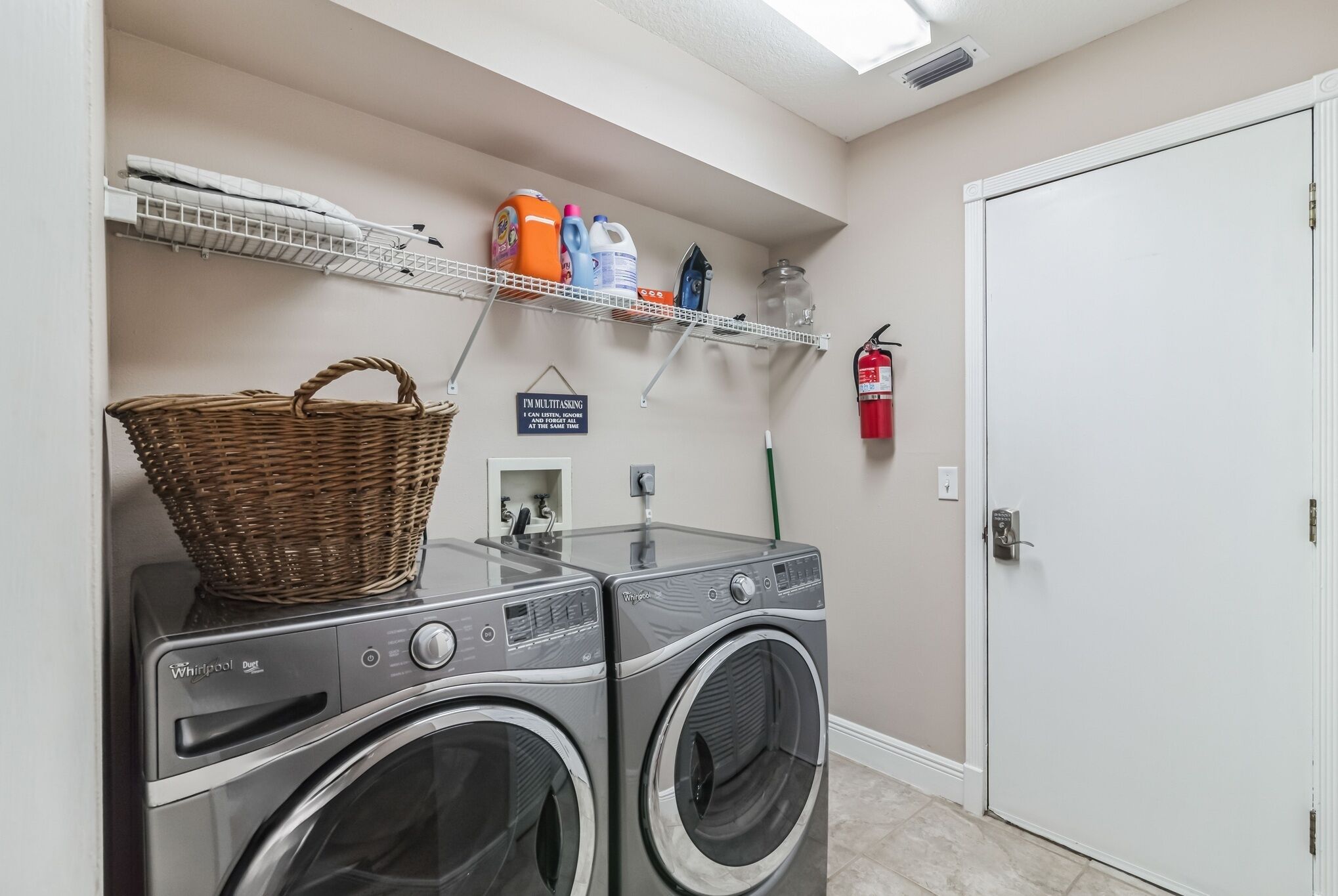 A laundry room with two front-loading Whirlpool dryers, a wicker basket on one dryer, and shelves above holding laundry supplies and a fire extinguisher.