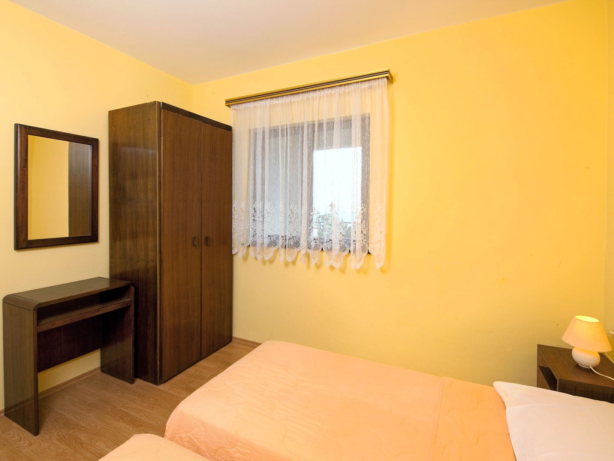 A bedroom with a bed, a wardrobe, a desk, a mirror, a window with sheer curtains, and a bedside lamp.