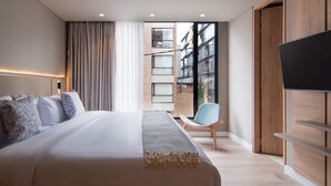 Ein modernes Hotelzimmer mit einem großen Bett, einem Fernseher, einem Sessel und Blick auf die Stadt durch ein großes Fenster.