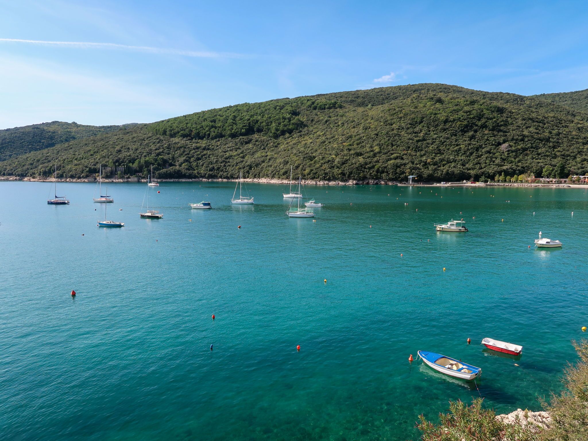 A coastal bay with multiple boats anchored in clear turquoise water, surrounded by lush green hills.