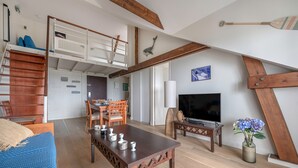 Un salon moderne avec un lit en mezzanine, des poutres en bois, un coin repas et un téléviseur.