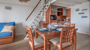 Un coin repas avec une table en bois dressée pour quatre personnes, un canapé bleu agrémenté d’un plaid beige et un escalier avec une rampe blanche menant à une cuisine.