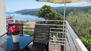 Un balcon doté d’une table, de chaises et d’un parasol, offrant une vue panoramique sur un plan d’eau et des collines.