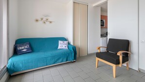 A room with a teal sofa, a black chair, and a kitchen in the background. - Seaview - flat for 4 (Pornichet)