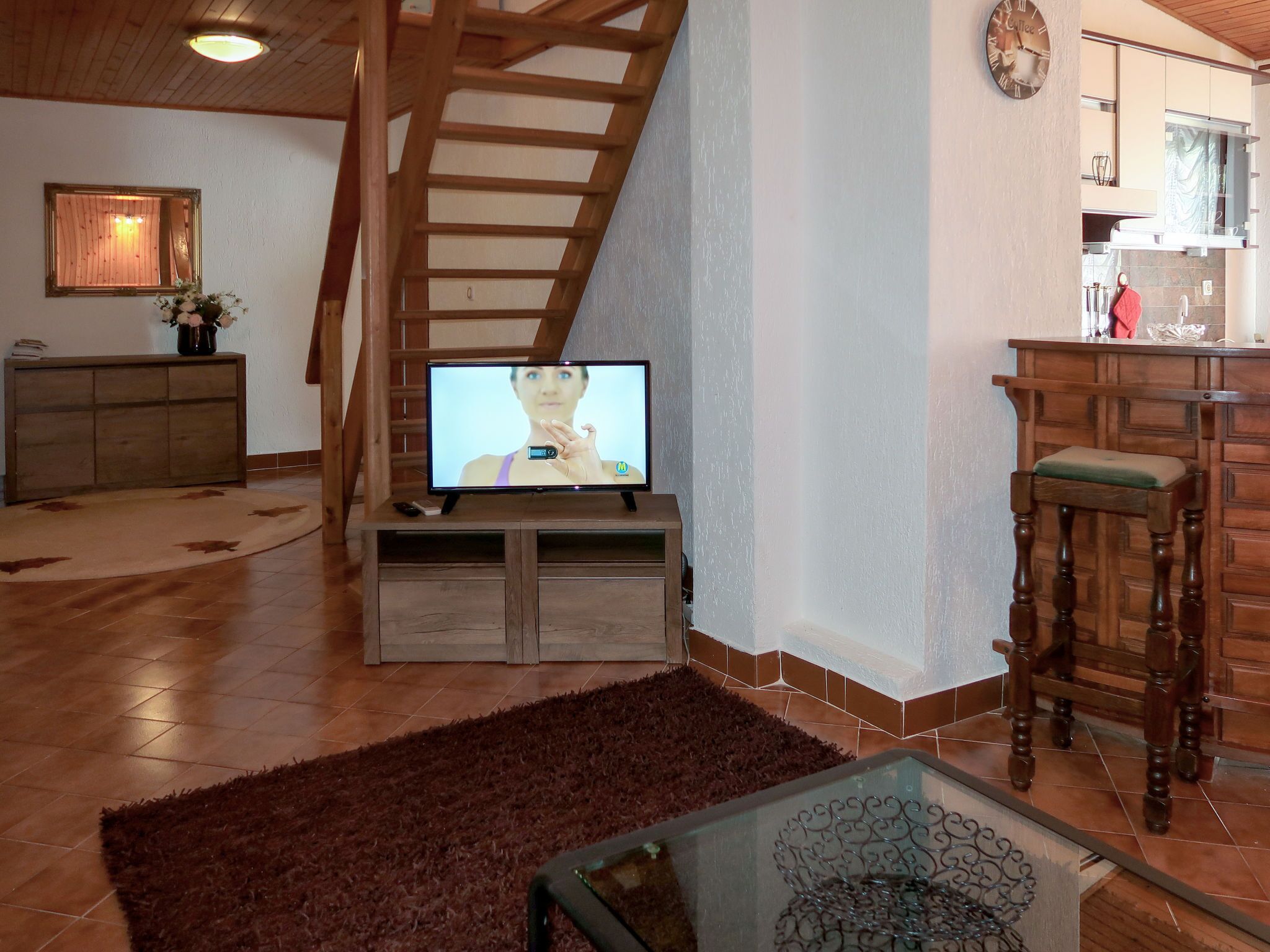 A living room with a wooden staircase, a television on a wooden stand, a kitchen area with a bar stool, and a decorative glass table.