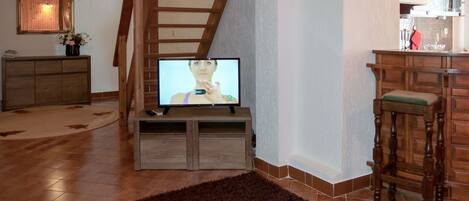 A living room with a wooden staircase, a television on a wooden stand, a kitchen area with a bar stool, and a decorative glass table.