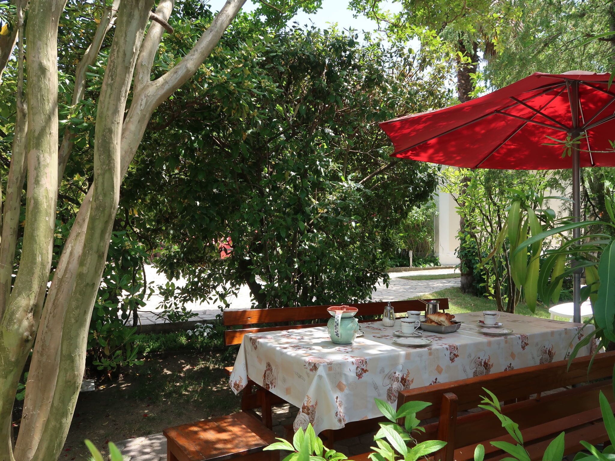 An outdoor dining area with a table set for two, surrounded by greenery and a red umbrella.
