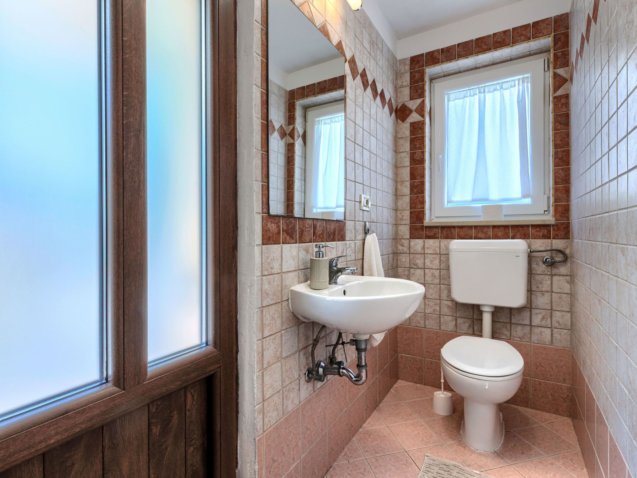 A bathroom with a wooden paneled door, a white sink, a toilet, and a window with a curtain.