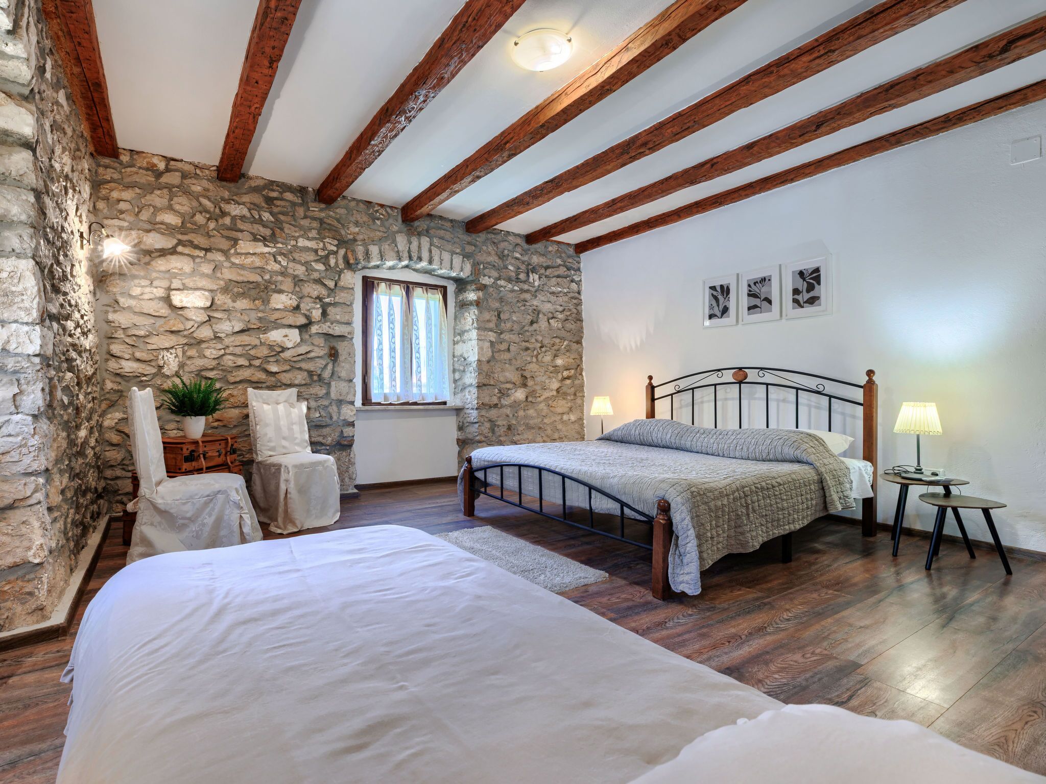 Une chambre avec un mur en pierre, des poutres en bois, un lit, une chaise, une petite table et une lampe.