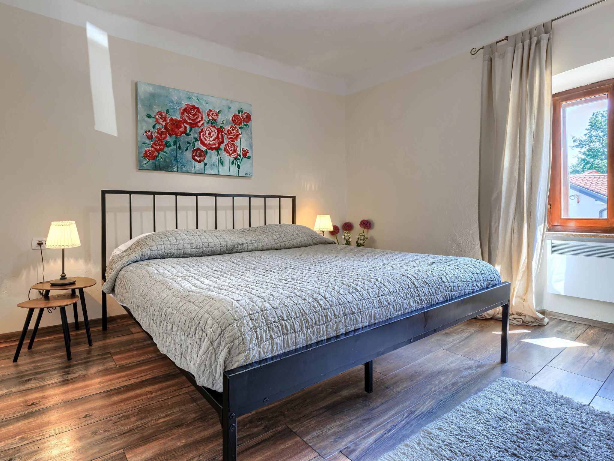 A bedroom with a bed, a nightstand, a lamp, a painting, and a window with curtains.
