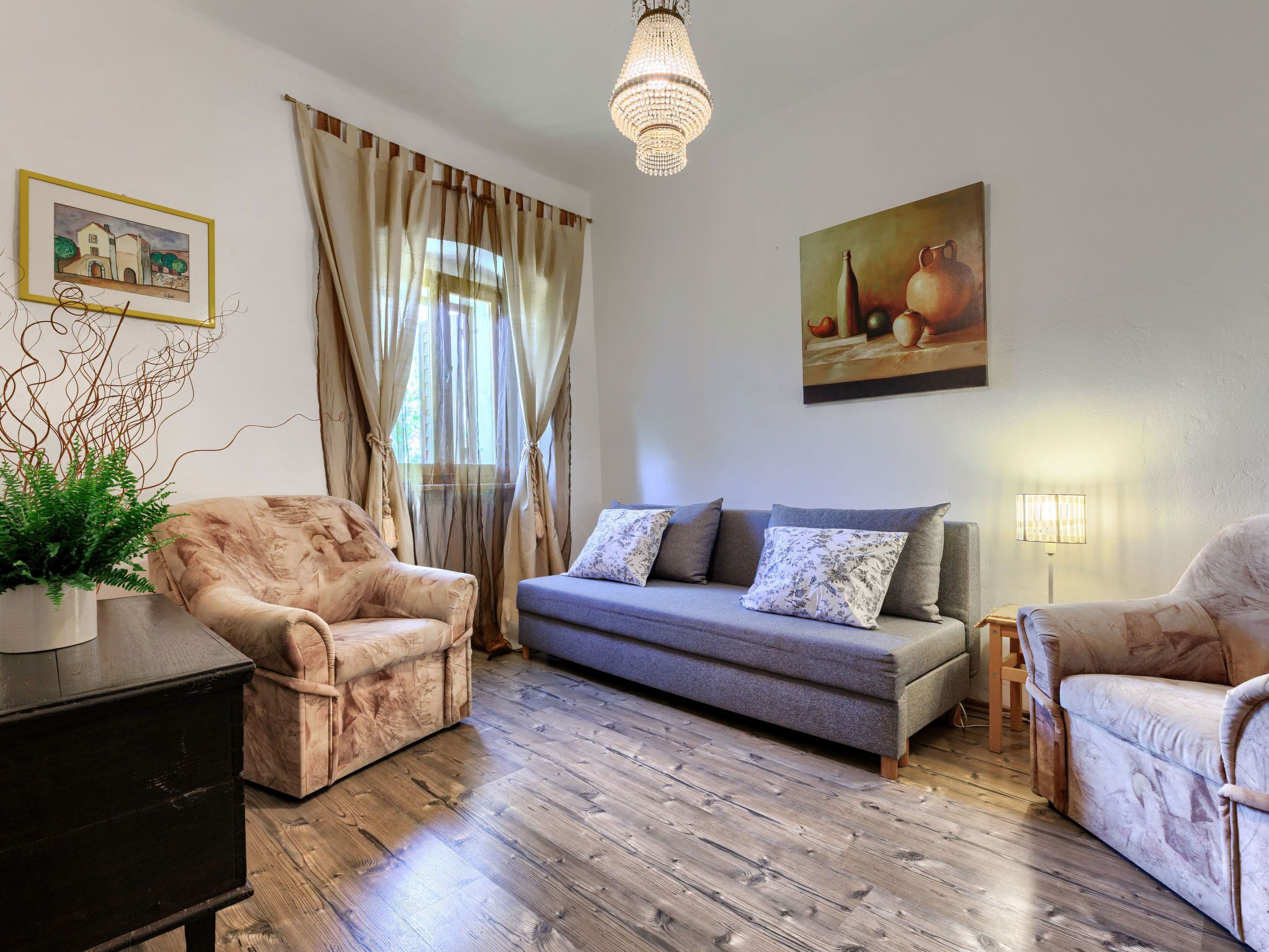 A living room with a sofa, two armchairs, a wooden floor, a chandelier, and a painting on the wall.