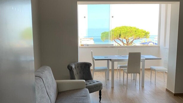 Una sala de estar moderna con un sofá gris, una mesa de comedor con sillas y un amplio ventanal con vista al mar.