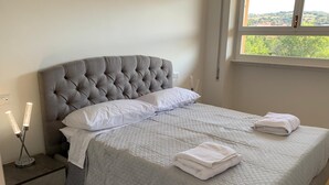 Una cama bien hecha con un cabecero gris capitonado, almohadas blancas y una colcha acolchada. Hay dos toallas dobladas sobre la cama.