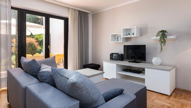 Ein modernes Wohnzimmer mit grauem Sofa, einem Flachbildfernseher, einer weißen Unterhaltungsecke und Blick ins Freie.
