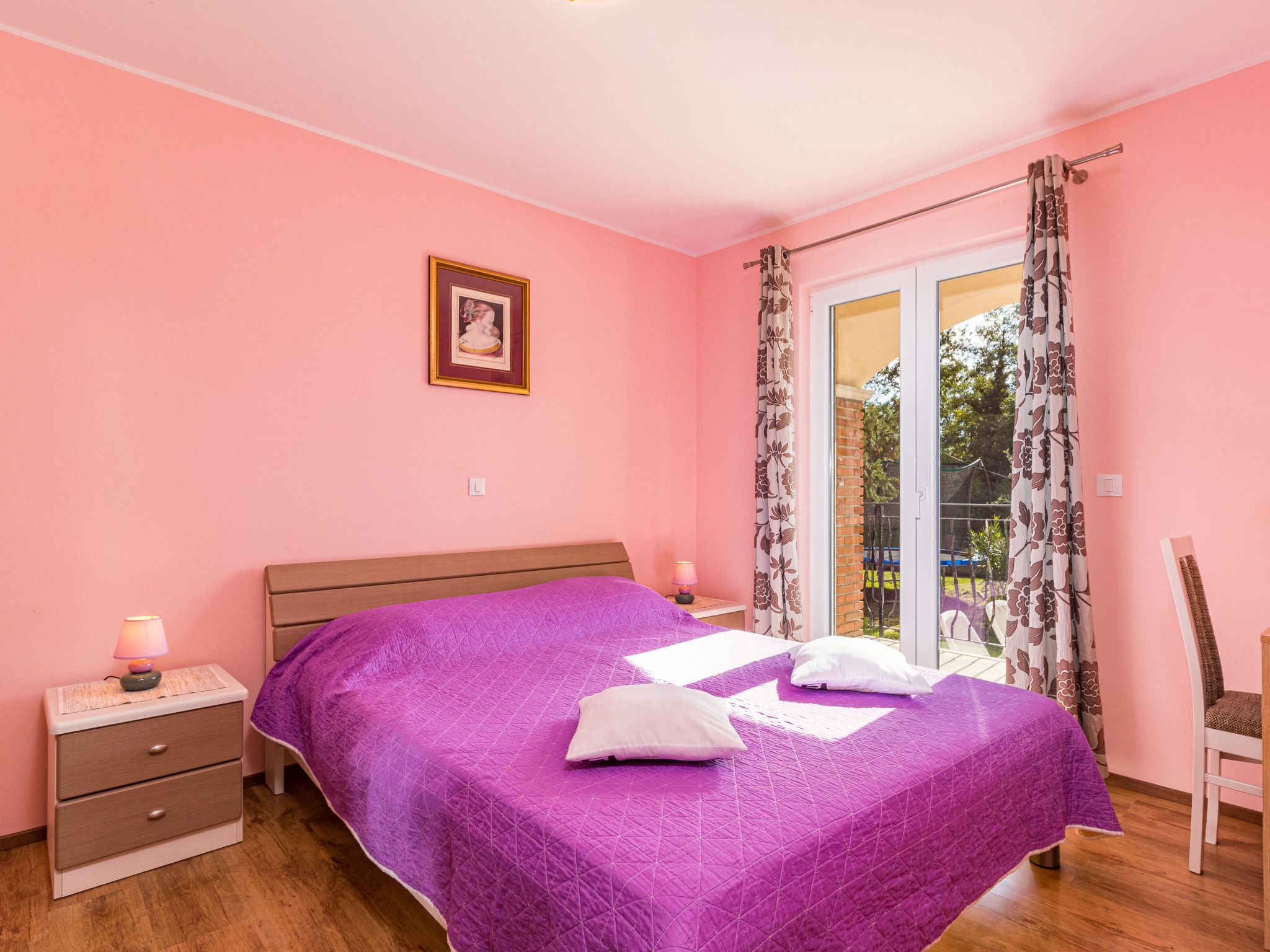 A bedroom with a bed, a nightstand, a chair, a window with curtains, and a picture on the wall.