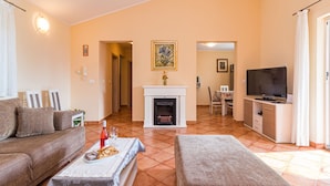 A living room with a sofa, ottoman, and a coffee table with a bottle and glasses. There is a fireplace, a TV, and a dining area in the background. - Vacation home Lara by Interhome (Poreč)