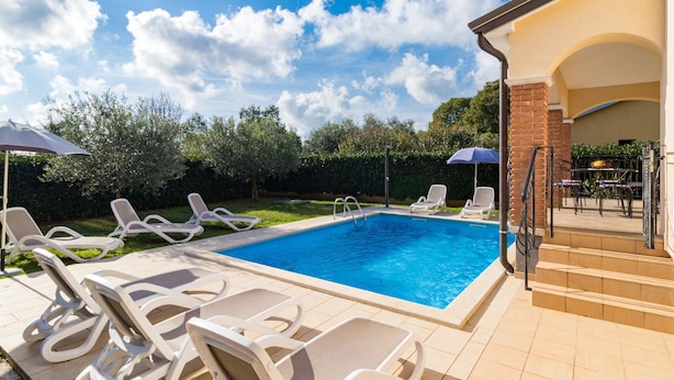 A rectangular swimming pool with white lounge chairs and umbrellas, surrounded by a brick wall and a garden with trees and shrubs. - Vacation home Lara by Interhome (Poreč)