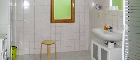 Un baño con baldosas blancas, un taburete de madera, un lavamanos con espejo y una ventana con marco verde.