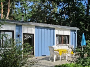 A small, single-story building with a blue exterior and a white door. There is a table with yellow tablecloth and white chairs outside. - Waldhäuser by Interhome (Himmelpfort)