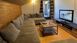 Ein gemütliches Wohnzimmer mit Holzboden, einer Couch, einem Couchtisch und einem Fernseher, der ein Radrennen zeigt.