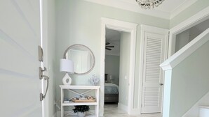Un pasillo con una puerta blanca, una consola blanca con espejo, una lámpara y una planta pequeña. Se ve una escalera a la derecha y una puerta que da a un dormitorio.