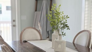 Una mesa de comedor con cubierta de madera oscura, sillas blancas y un camino de mesa blanco. Encima de la mesa, hay una repisa metálica con frascos de vidrio y una planta en maceta.