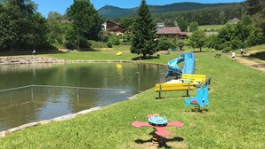 Un parque con un estanque, juegos infantiles y casas al fondo.