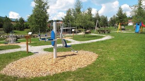 Un parque infantil con varios juegos, incluyendo columpios y un tobogán, rodeado de árboles y un sendero de grava.