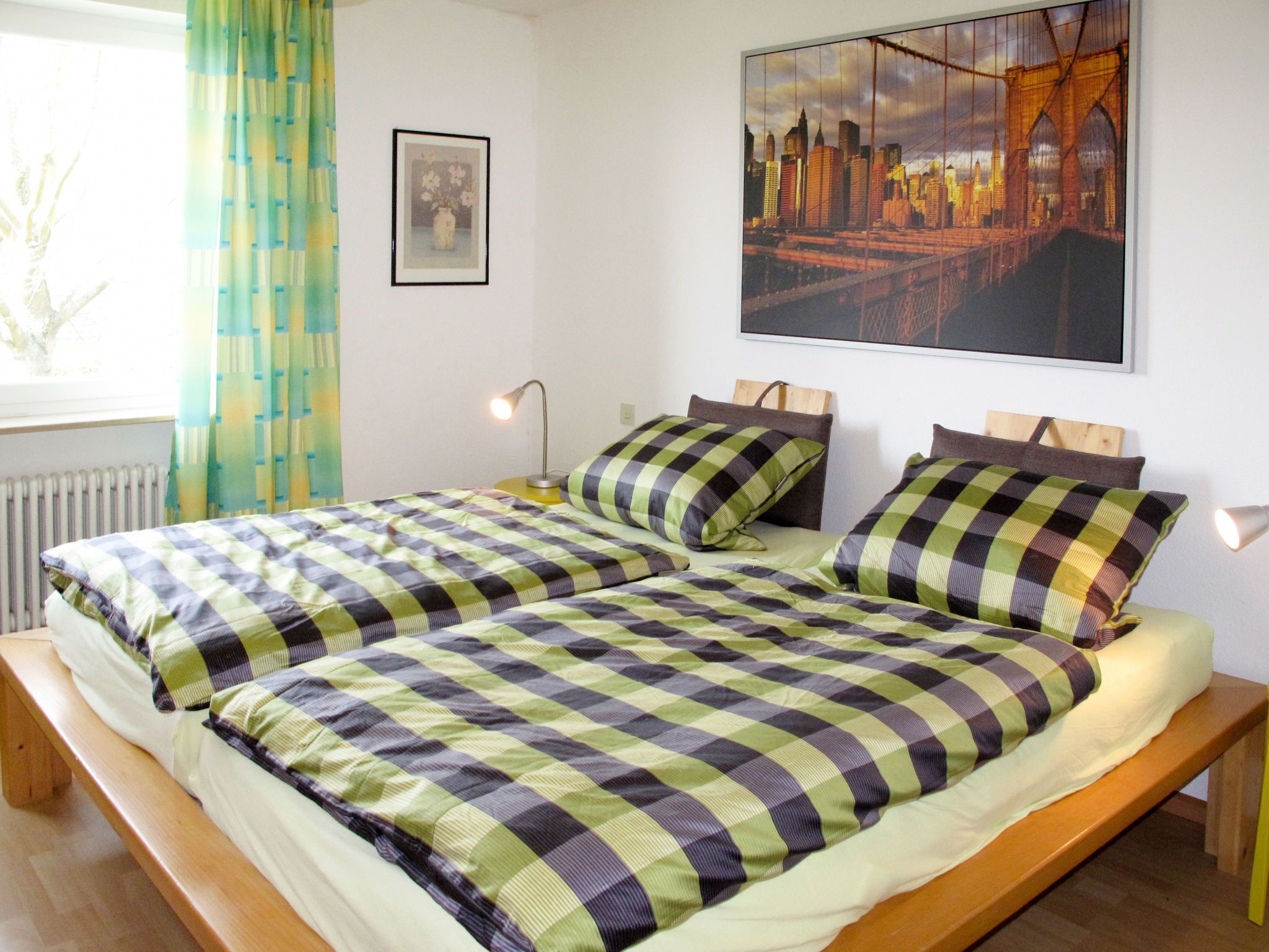 A double bed with a checkered bedspread, a bedside table with a lamp, a framed picture on the wall, and a large cityscape painting above the bed.