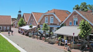 Calle bordeada por edificios tradicionales de ladrillo, algunos con tejados a dos aguas y otros planos. Hay zonas de estar al aire libre con mesas y sillas, y se ven algunas personas caminando o sentadas.