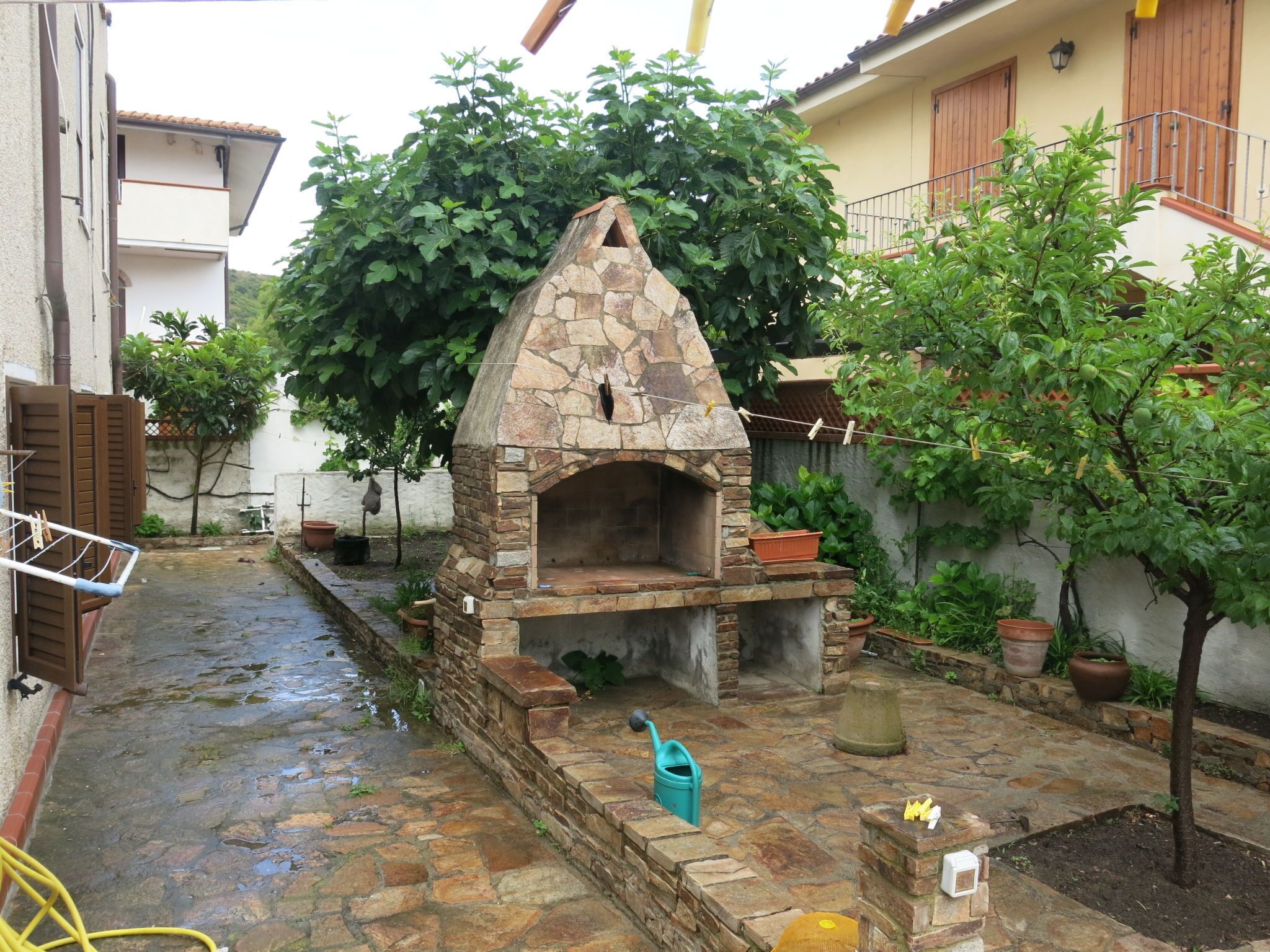 Un área de cocina exterior de piedra con chimenea, rodeada de plantas en macetas y un pequeño árbol.